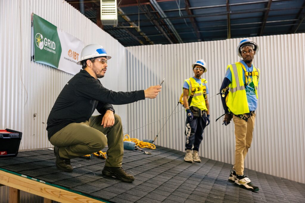 An instructor kneels on a practice roof deck demonstrating a technique to two trainees in high-visibility vests.