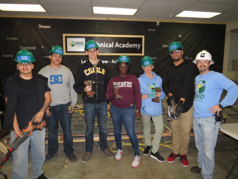 Seven people wearing hard hats pose together at a Technical Academy event, holding tools and wearing GRID-branded apparel