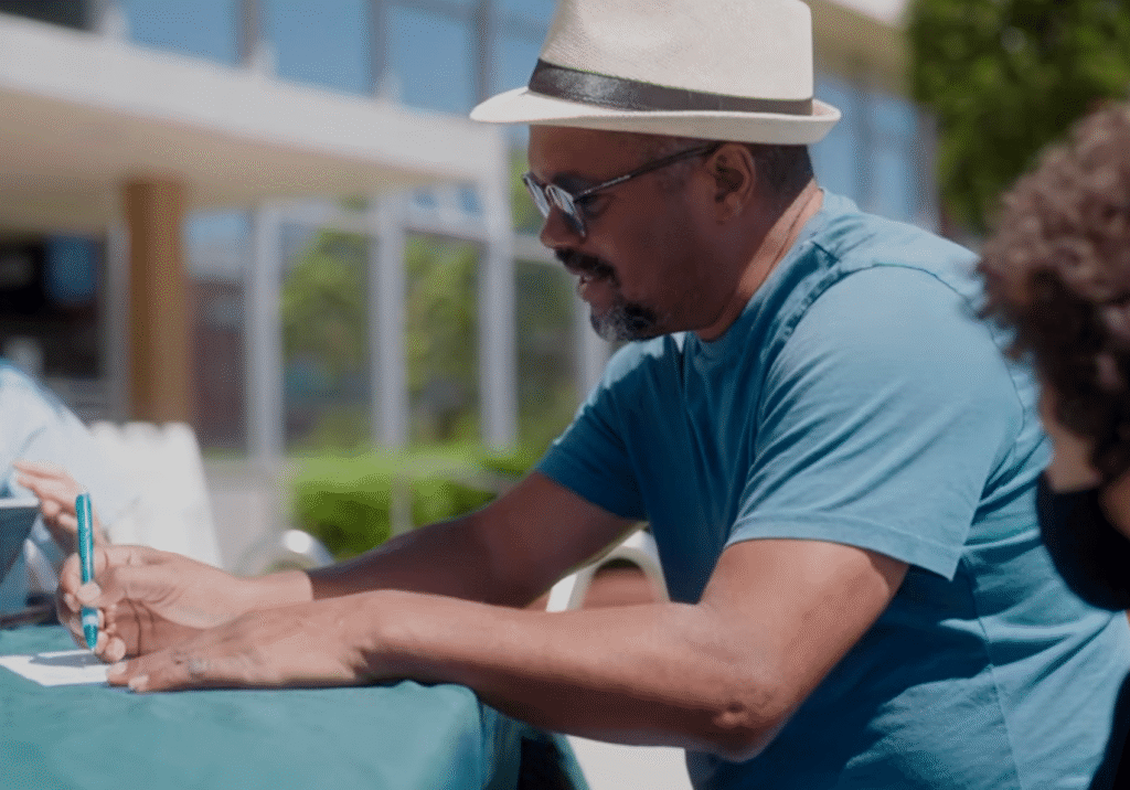 A man in a sun hat and glasses signs a form at an outdoor table.