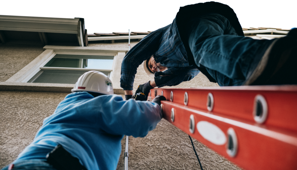 Two installers pass a ladder up the side of a building from a low, dramatic angle.