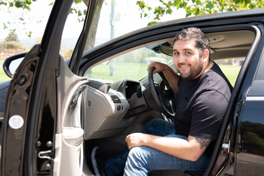 A man sits in the driver's seat of an electric vehicle with the door open, smiling at the camera.