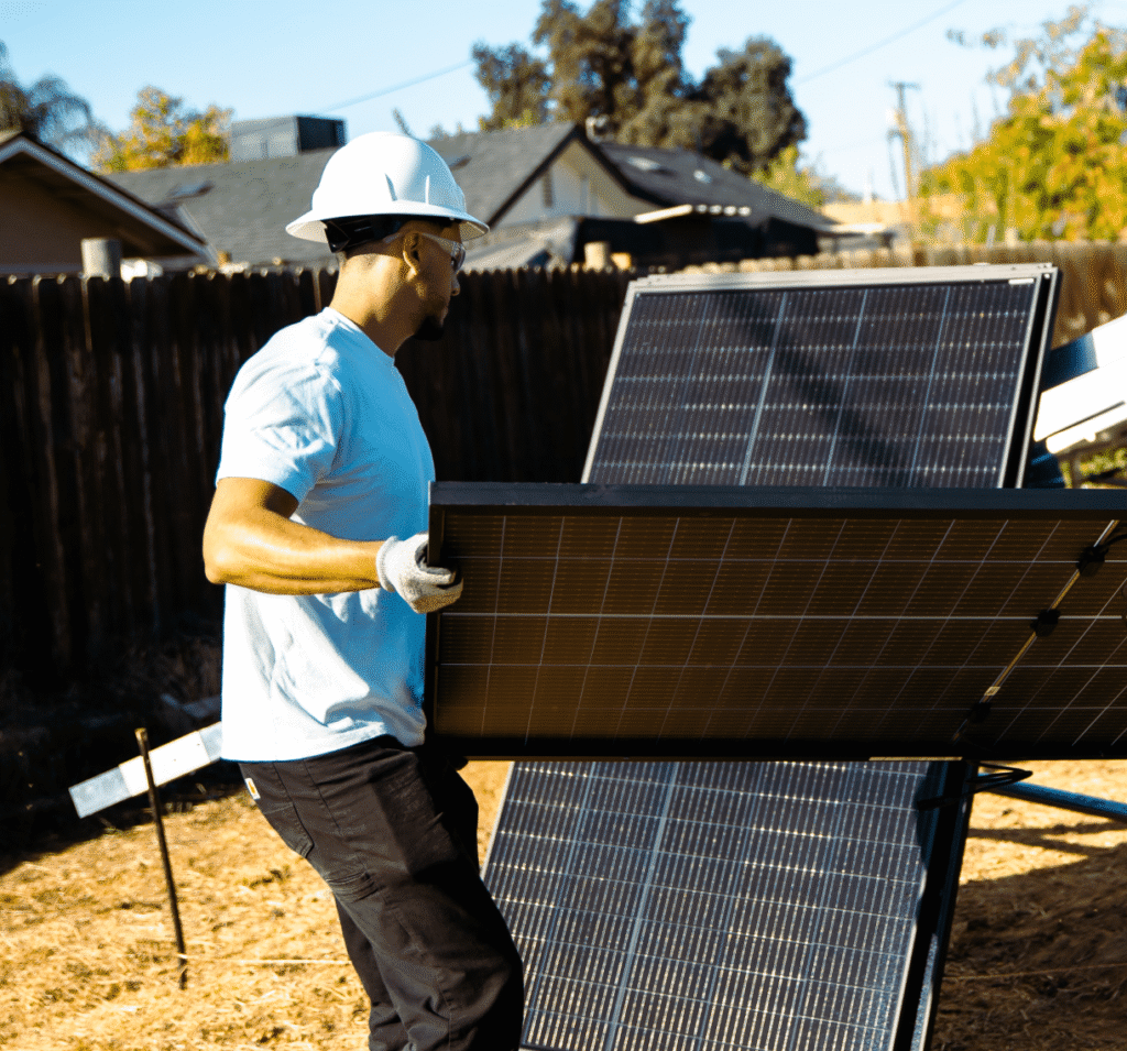 A man carries a battery storage unit across a yard toward a home.
