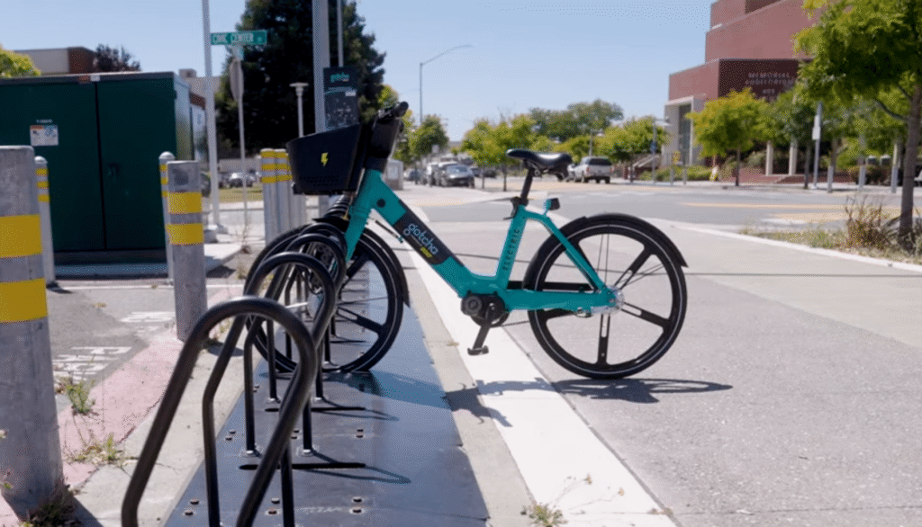 A teal electric bicycle from a bike-share program sits parked at a downtown docking station.