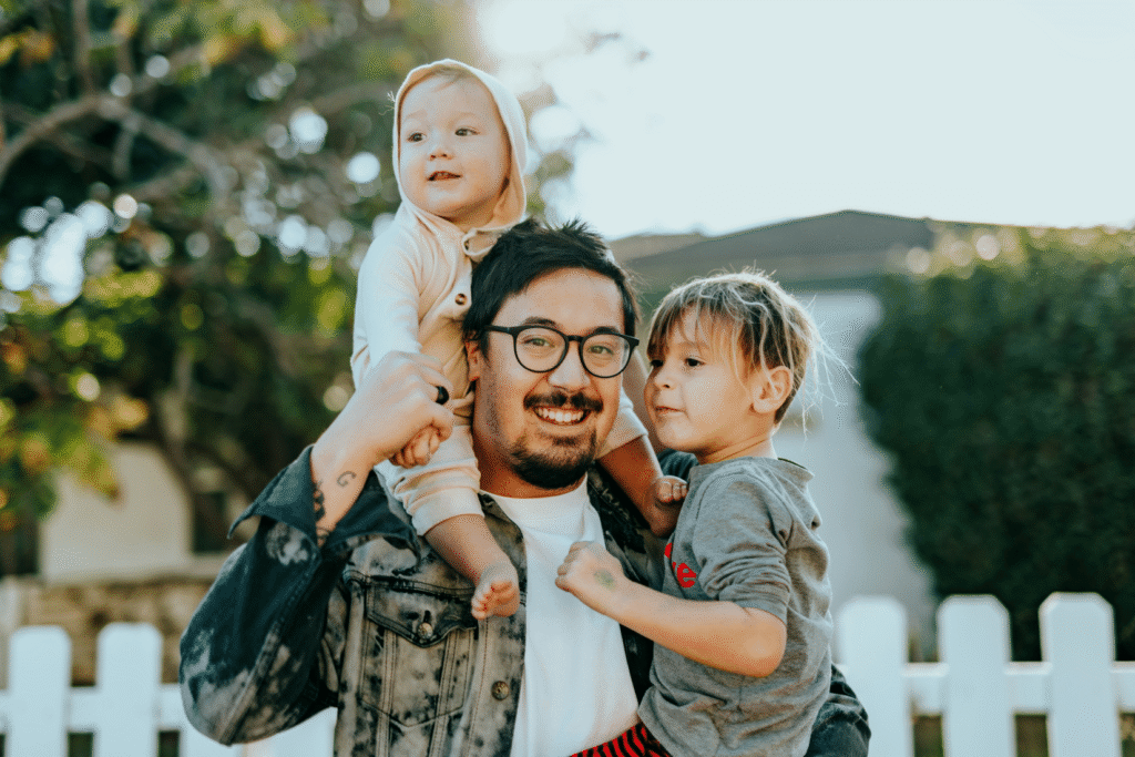 A father carries two children on his shoulders in a backyard.
