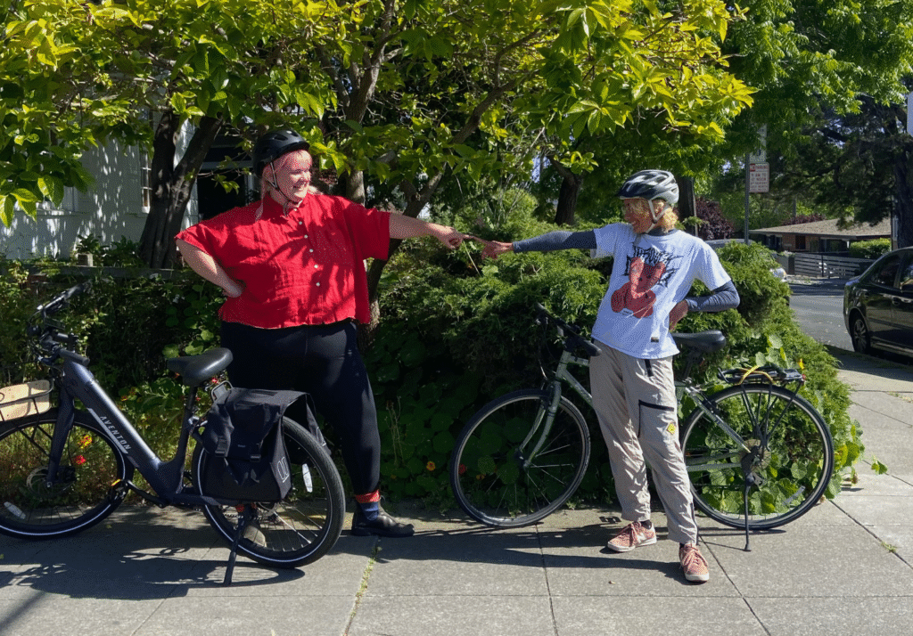Two cyclists in helmets stand with their bikes on a sidewalk, reaching out to fist-bump.