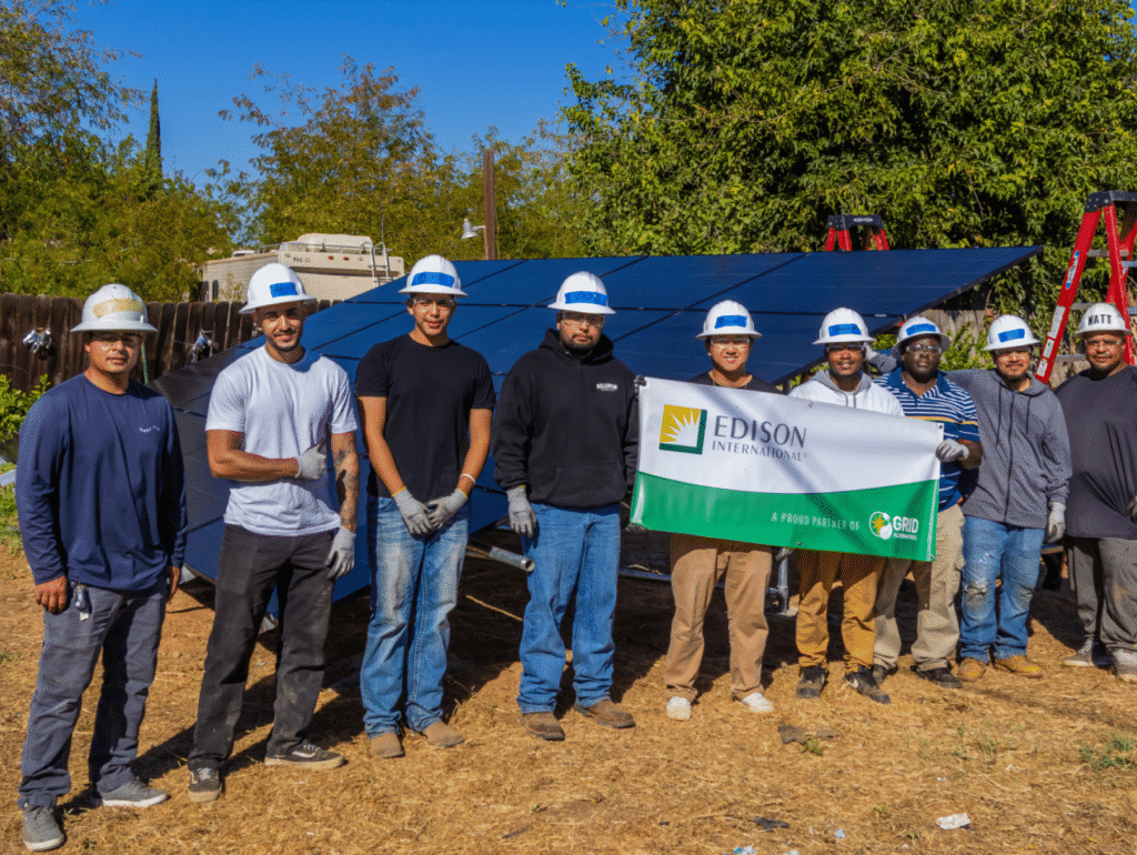 Nine installers and volunteers in hard hats standing behind a ground-mounted solar array, two of them holding an Edison International banner reading A proud partner of GRID Alternatives.