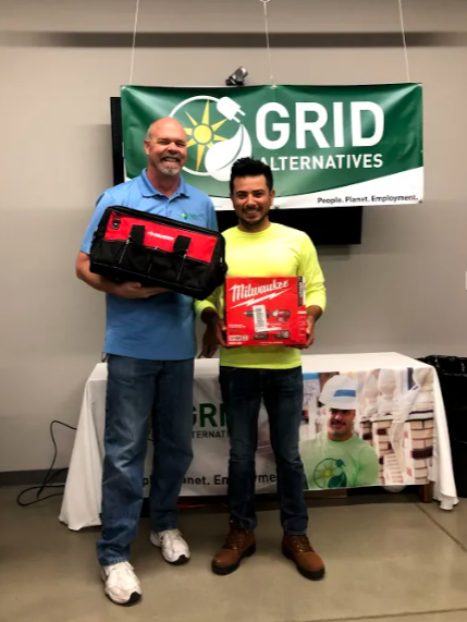Two men holding red Milwaukee tool boxes in front of GRID Alternatives banner