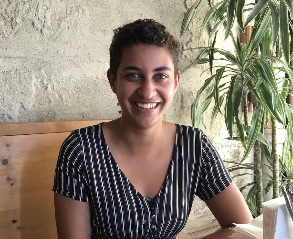 Smiling person wearing black striped shirt, sitting indoors with potted plant and textured wall behind them