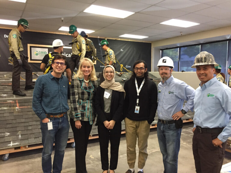 Six people posing at a GRID construction safety training event with instructors demonstrating techniques on an elevated platform behind them