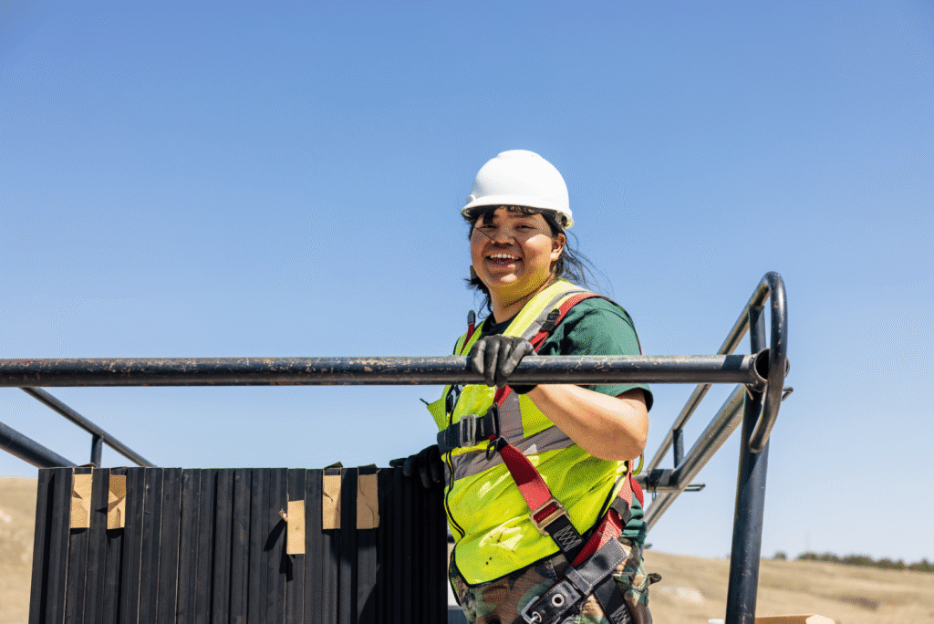 A smiling installer in a hard hat and safety harness holding a scaffold rail on a rooftop, dry hills in the background.