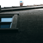 A worker in a white hard hat looks out over the ridge of a house roof, seen from the ground below.
