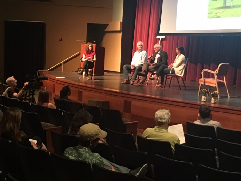 Panel discussion on stage with four speakers seated in chairs and audience members in foreground, one operating video camera