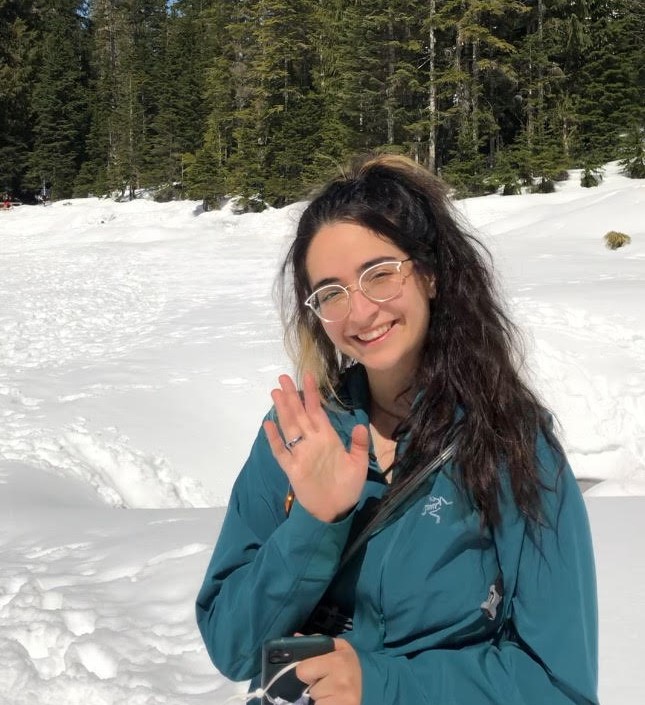 Shadeh waves and smiles at camera, wearing a teal windbreaker, clear glasses, and crossbody purse. They stand on freshly fallen snow with lush pine trees and a clear blue sky in the background.