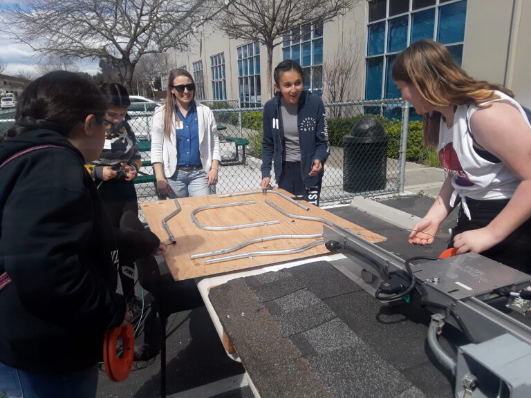 Five students examine a wooden circuit board model outdoors in a courtyard setting