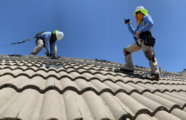 Two workers with safety gear performing roof tile installation on a steep residential roof under clear sky