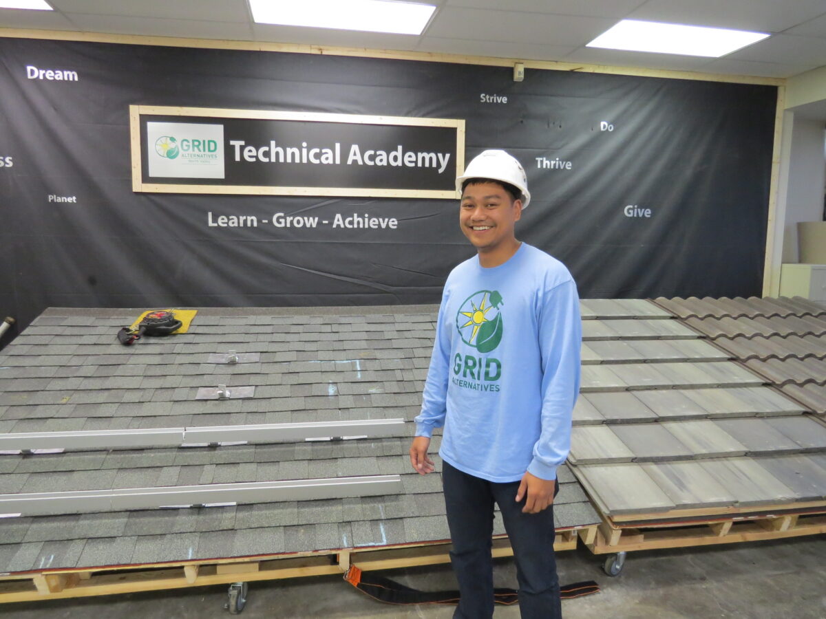 Student in GRID Alternatives shirt and hard hat standing in Technical Academy with roofing materials displayed
