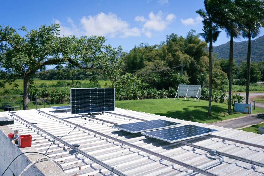 Rows of solar panels mounted on a low roof, with palm trees and hills behind.