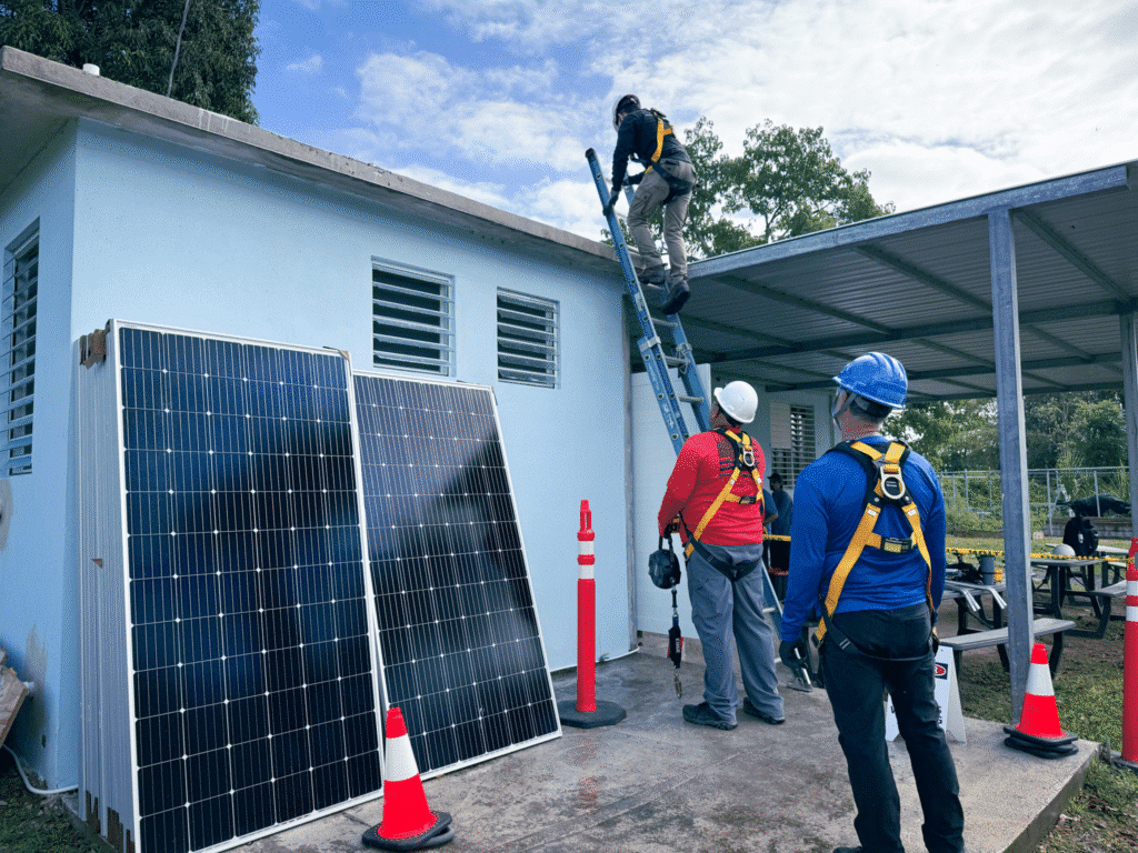 Installers in hard hats carry a solar panel past a carport frame on site.