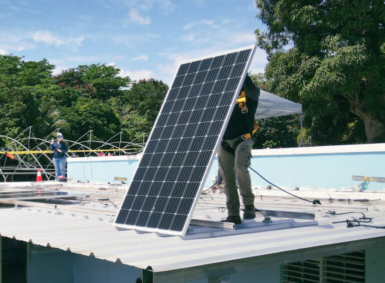 Worker installing large solar panel on flat roof with trees and blue sky in background