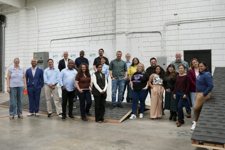 Group of approximately 25 people posing together in an industrial warehouse space with white brick walls and exposed pipes