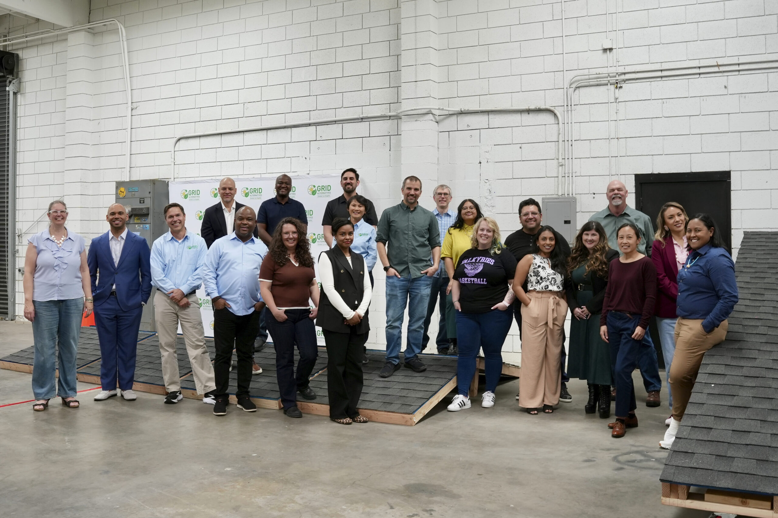 Group of approximately 25 people posing together in an industrial warehouse space with white brick walls and exposed pipes