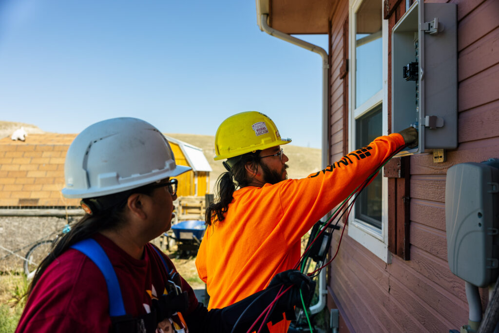 Two construction workers in safety gear installing electrical equipment on a brick building exterior