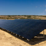 Large solar panel array installed on sandy ground with rolling hills and trees in the background under blue sky