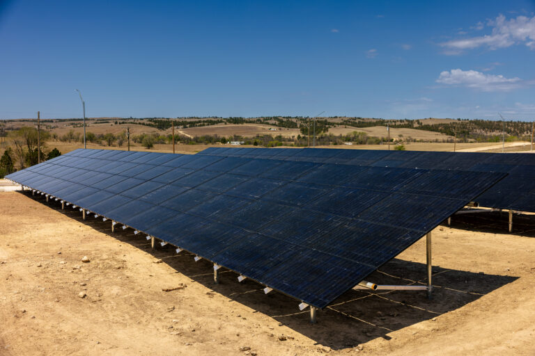 Large solar panel array installed on sandy ground with rolling hills and trees in the background under blue sky
