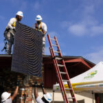 Workers installing solar panel on residential roof with ladder and safety equipment