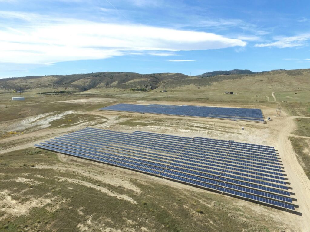 Aerial view of two large solar panel arrays on dusty terrain with rolling hills and grassland in background