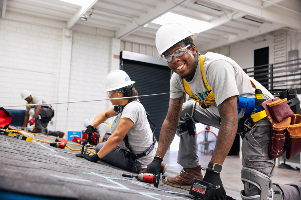 Construction workers in hard hats and safety gear practicing roofing techniques on a training surface indoors