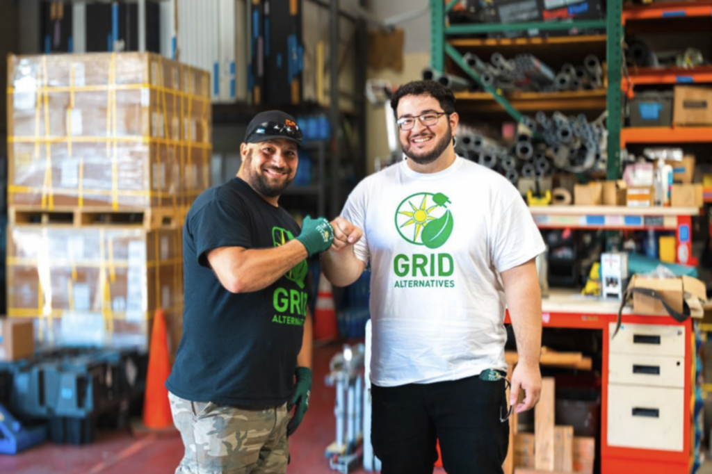 Two GRID Alternatives employees fist bumping in a warehouse with shelving and equipment behind them