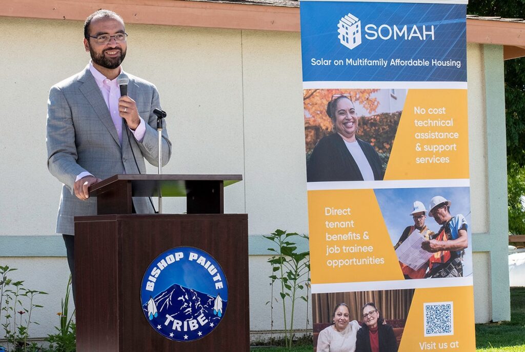 Man speaking at podium with Bishop Paiute Tribe logo, presenting SOMAH solar housing program information on display board
