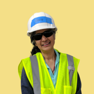 Woman wearing hard hat with "Vance" written on blue band, safety vest, and sunglasses against yellow background