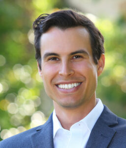 Professional headshot of smiling man in navy blazer and white shirt against blurred green background