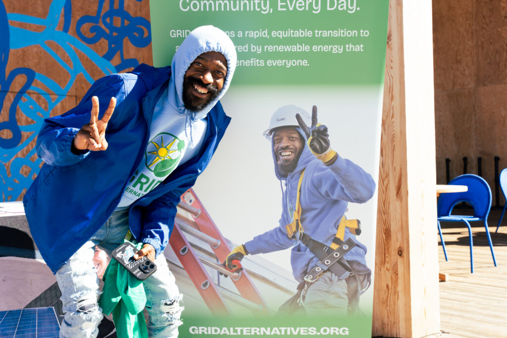 Man in blue jacket posing with peace sign in front of GRID Alternatives renewable energy community banner