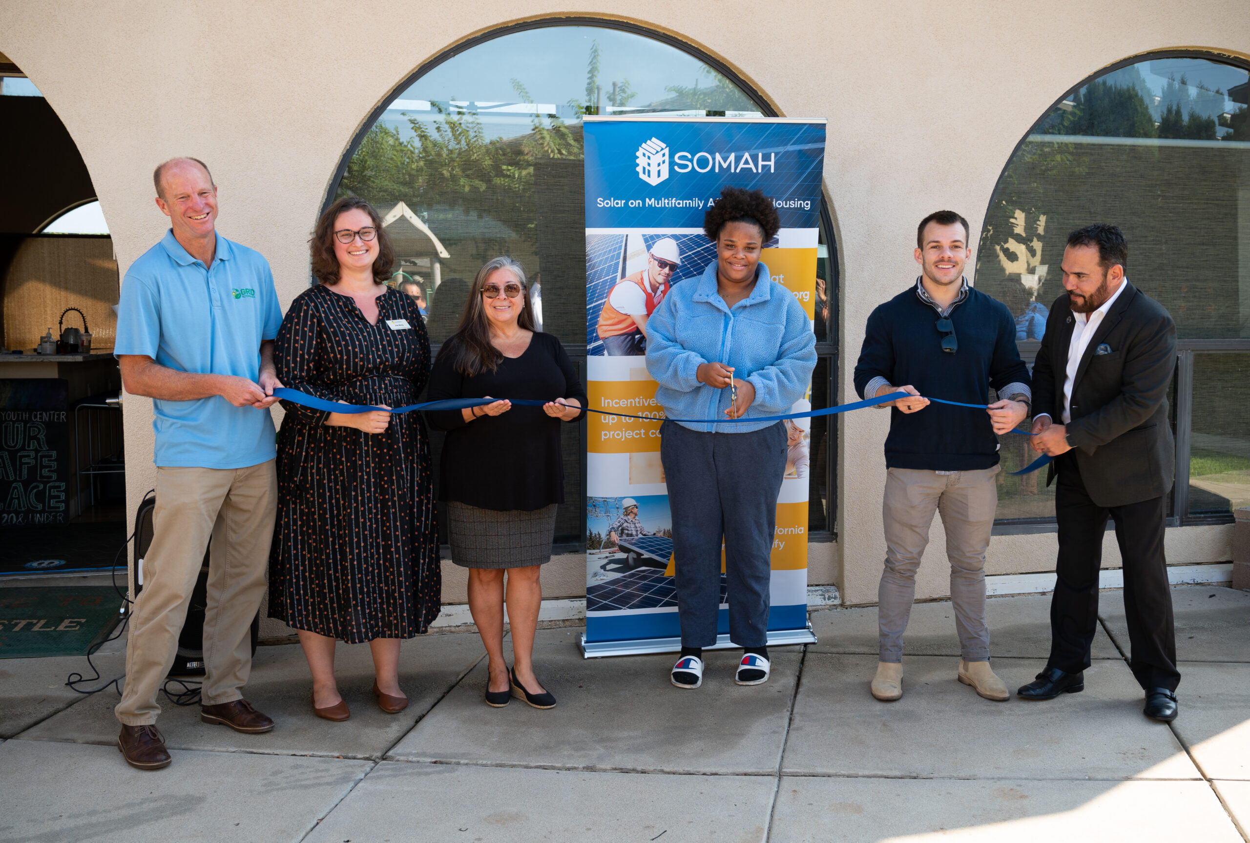 Six people cutting a blue ribbon at a SOMAH solar housing project ribbon-cutting ceremony