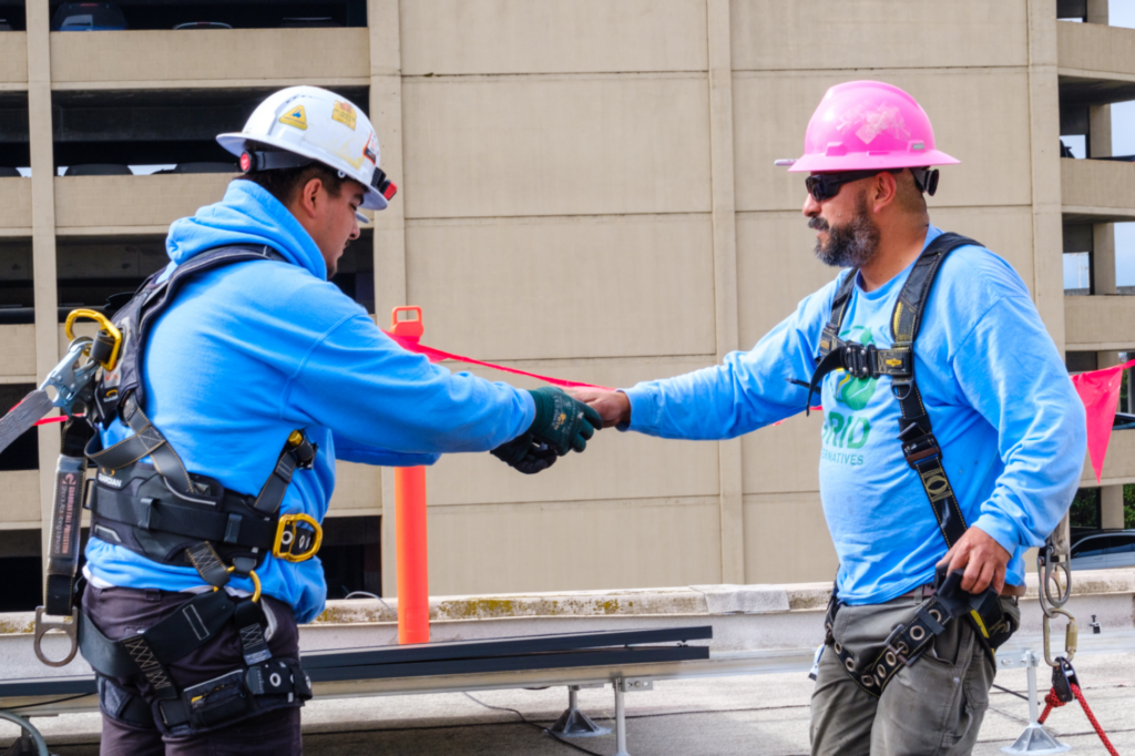 Two GRID installers in hard hats shake hands on a job site beside a partly built solar array