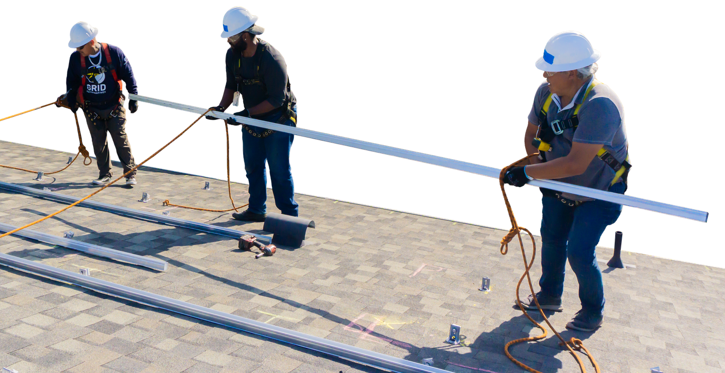 Three installers in hard hats and harnesses passing a length of racking rail across a shingle roof.