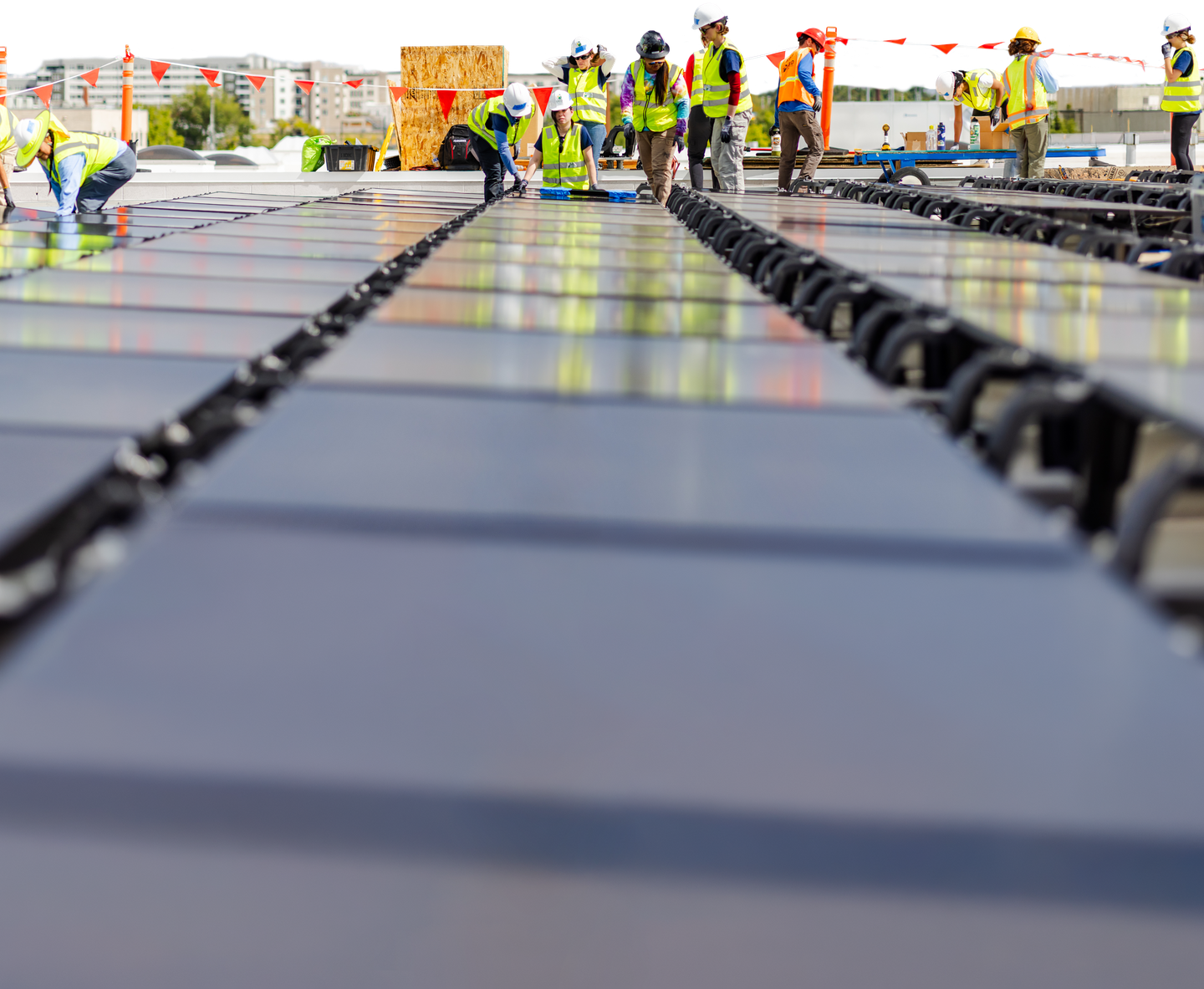 A crew of installers in hi-vis vests and hard hats working across rows of solar panels on a flat commercial roof, city skyline behind them.