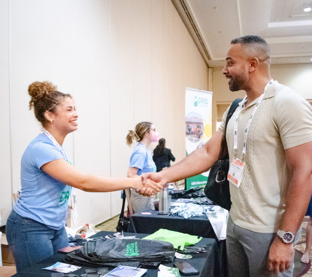 Two people shaking hands at a conference booth displaying promotional materials and merchandise on a black table
