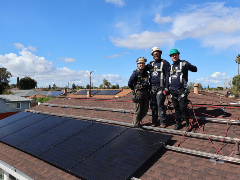 Three solar installation workers in safety gear stand on a residential rooftop with solar panels and blue sky