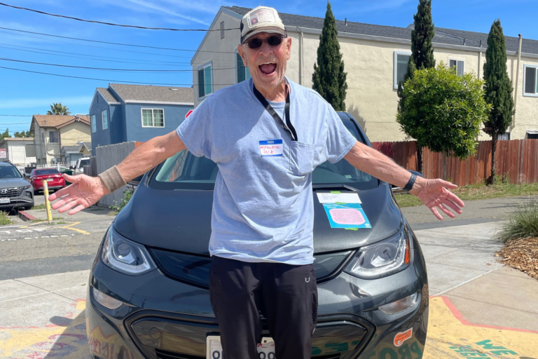 Man in white cap and shirt standing in front of black electric car with arms outstretched, smiling at camera on residential street