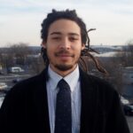 Portrait of Julian, a man with dreadlocks wearing a black blazer and checkered shirt with tie, outdoors with cityscape background